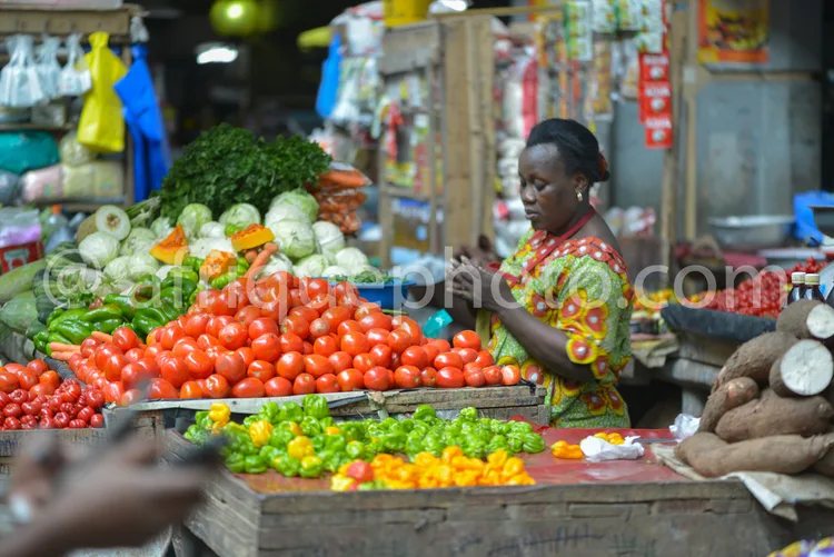 Africa photo — Une femme souriante, vêtue d’un habit traditionnel, se tient derrière un étalage rempli de légumes variés : tomates rouges, chou vert,