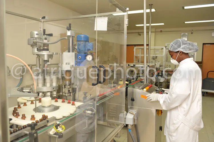 Africa photo — Un technicien en combinaison stérile blanche, masque et charlotte, manipule une machine industrielle de remplissage de flacons médicaux