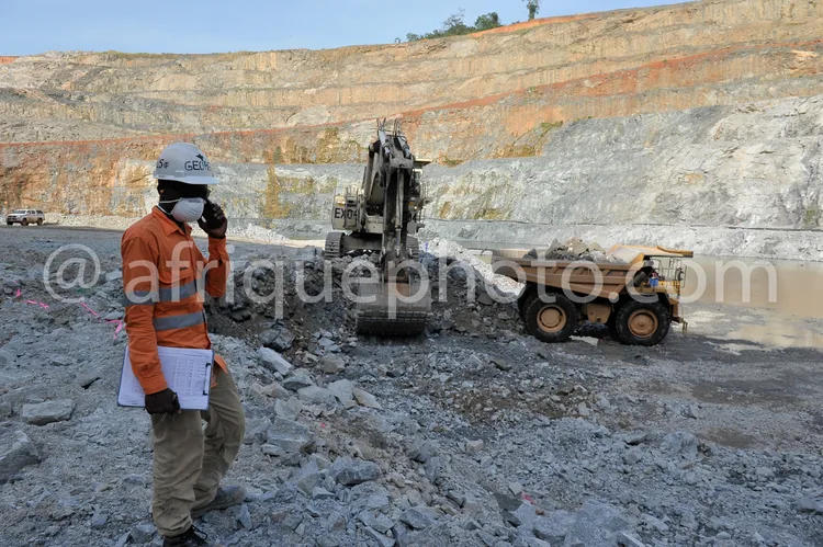 Africa photo — Un superviseur de mine équipé d’un casque blanc et d’une veste orange, tenant un carnet et un téléphone, se tient près d’un engin de ch