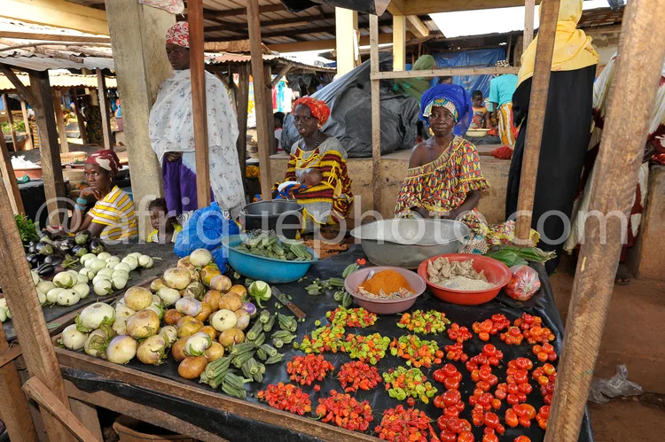 Africa photo — Image d’un étal de marché traditionnel où trois femmes, vêtues de tenues colorées et de foulards sur la tête, vendent des légumes frais