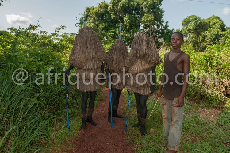 Africa photo — Un homme souriant en tenue simple se tient à côté de trois mannequins habillés de costumes traditionnels en paille, alignés sur un chem