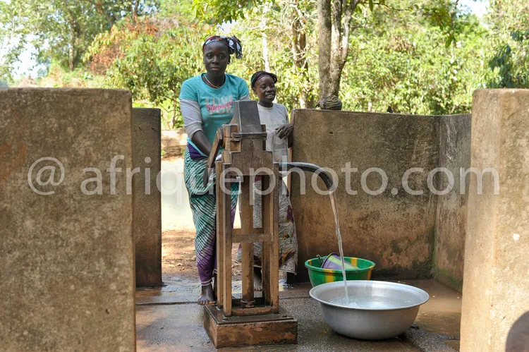 Africa photo — Deux femmes souriantes actionnant une pompe à eau en bois près d'un puits, avec un seau métallique rempli d'eau sous le jet, dans un ca
