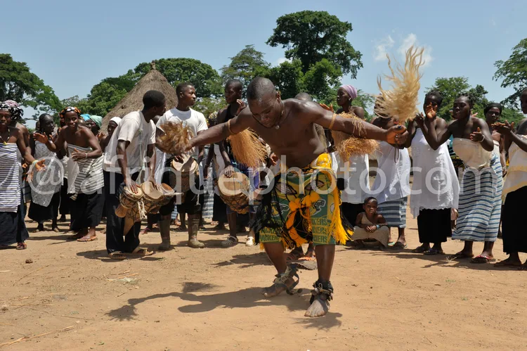 Africa photo — Un groupe de personnes participe à une danse traditionnelle africaine en extérieur. Un homme torse nu, vêtu d’un pagne jaune et noir, l