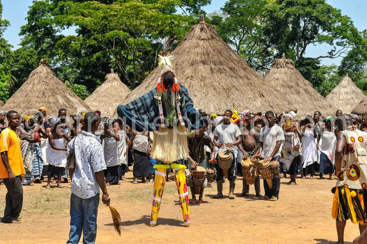 Africa photo — Un danseur traditionnel vêtu d’un costume coloré et masqué exécute une danse au centre d’une assemblée. Autour de lui, des spectateurs