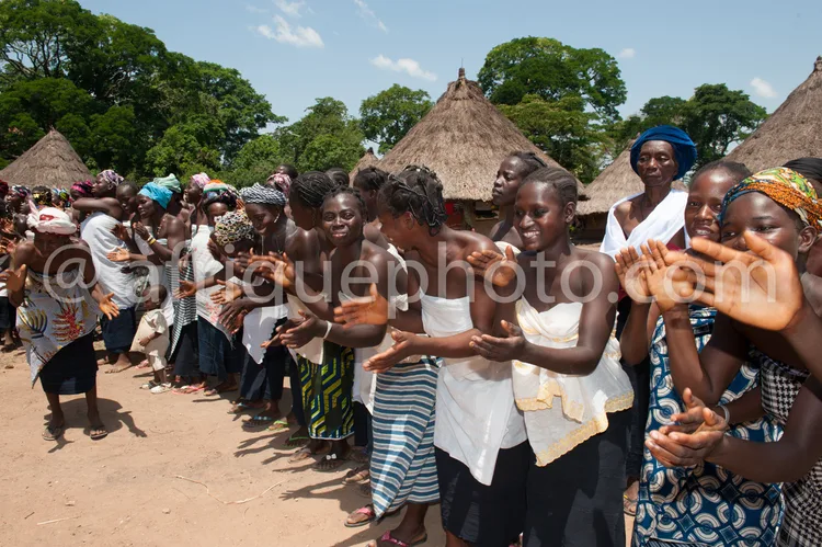 Africa photo — Groupe de femmes souriantes, vêtues de tenues colorées et légères, participant à une danse collective en plein air devant des habitatio