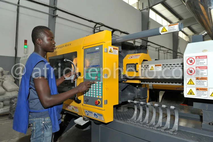Africa photo — Un opérateur en bleu, debout devant une machine CNC jaune et grise, interagit avec un écran tactile pour contrôler l'usinage de pièces