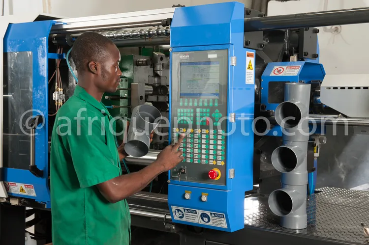 Africa photo — Un technicien en tenue verte se tient devant une machine industrielle bleue et grise, appuyant sur des boutons d’un écran tactile pour