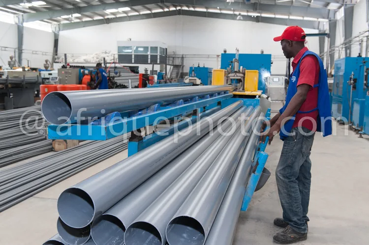 Africa photo — Un ouvrier en tenue de travail rouge et casquette rouge examine attentivement une série de tubes métalliques gris posés sur une table d