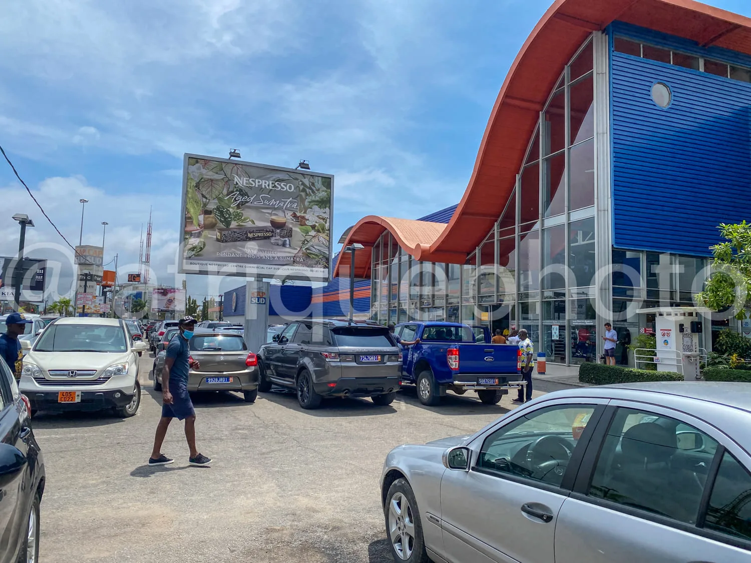 Africa photo — Image montrant une boutique Nespresso située devant un centre commercial moderne aux couleurs bleues et orange, avec une file de voitur
