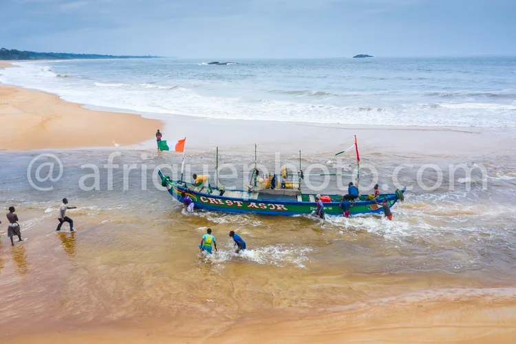 Africa photo — Un groupe de pêcheurs aide à guider un bateau peint en bleu et vert, nommé 'Crist ASOR', vers la plage. Les vagues léchent le sable fin