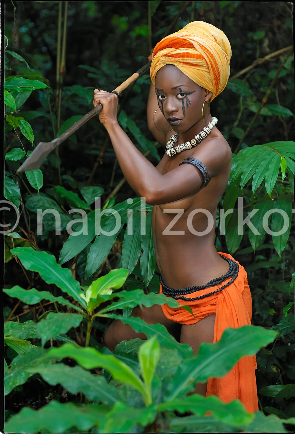 Africa photo — Une jeune femme se tient dans une forêt luxuriante, vêtue d’une tenue orange et d’un turban aux reflets dorés. Elle porte des accessoir