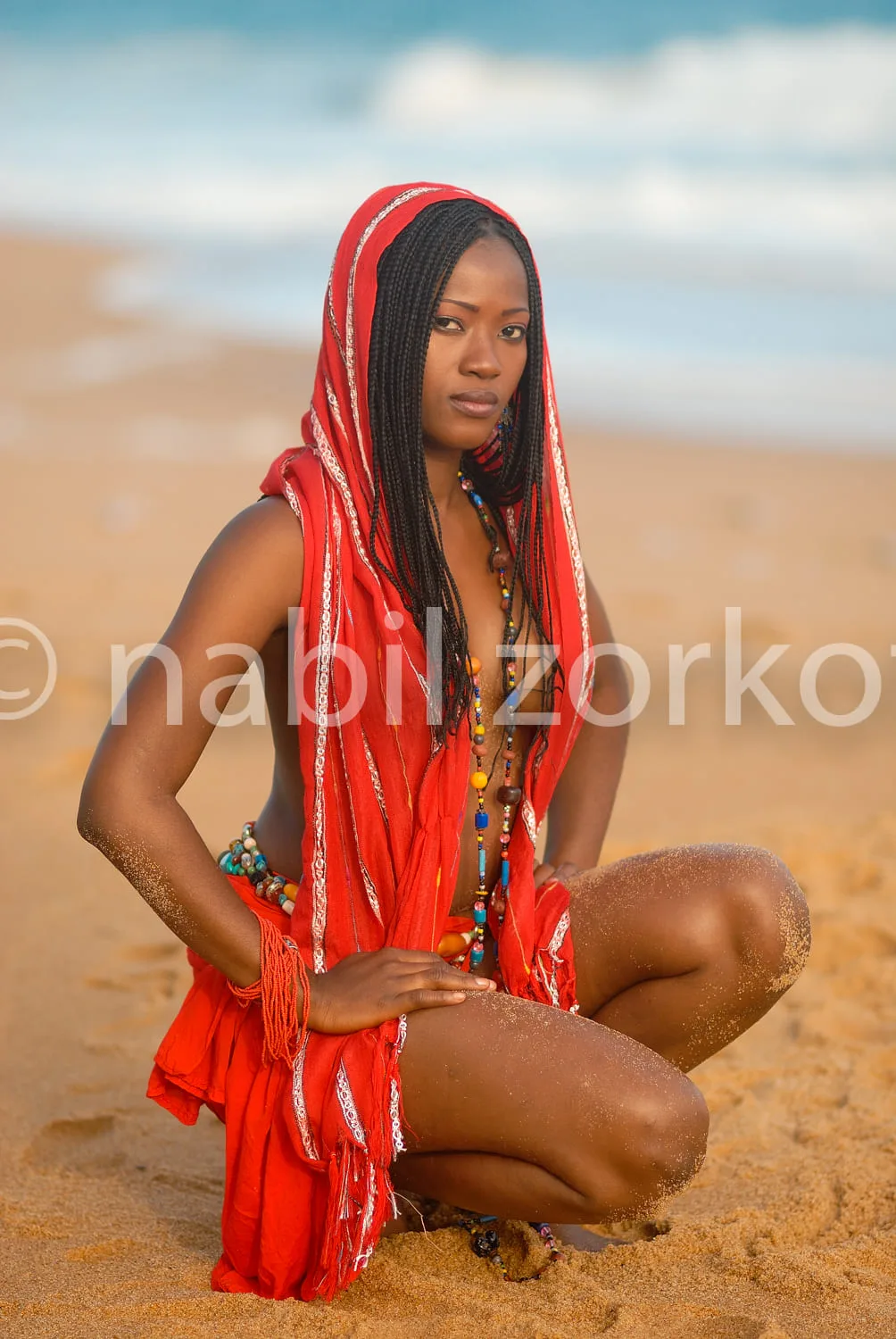 Africa photo — Une femme assise sur le sable, vêtue d'une robe rouge traditionnelle à motifs brodés, coiffée de tresses et parée de multiples colliers