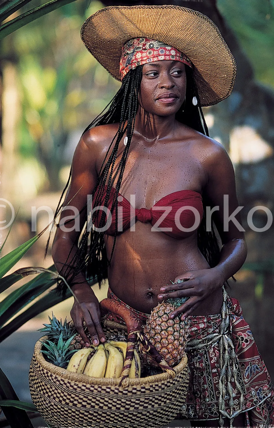 Africa photo — Une femme porte un panier rempli de bananes et d’ananas, vêtue d’un pagne coloré et d’un chapeau en paille. Ses cheveux sont tressés et