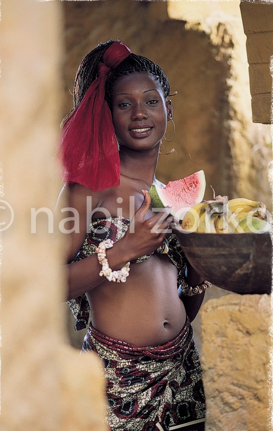 Africa photo — Une jeune femme souriante, vêtue d'une tenue traditionnelle colorée avec un pagne imprimé et un voile rouge, tient un morceau de pastèq