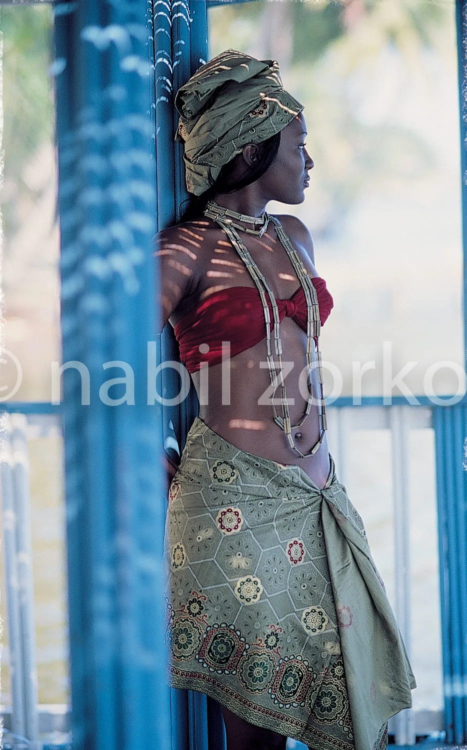 Africa photo — Une femme debout près d'une structure bleue, vêtue d'une tenue traditionnelle composée d'un haut rouge sans manches, d'une jupe longue
