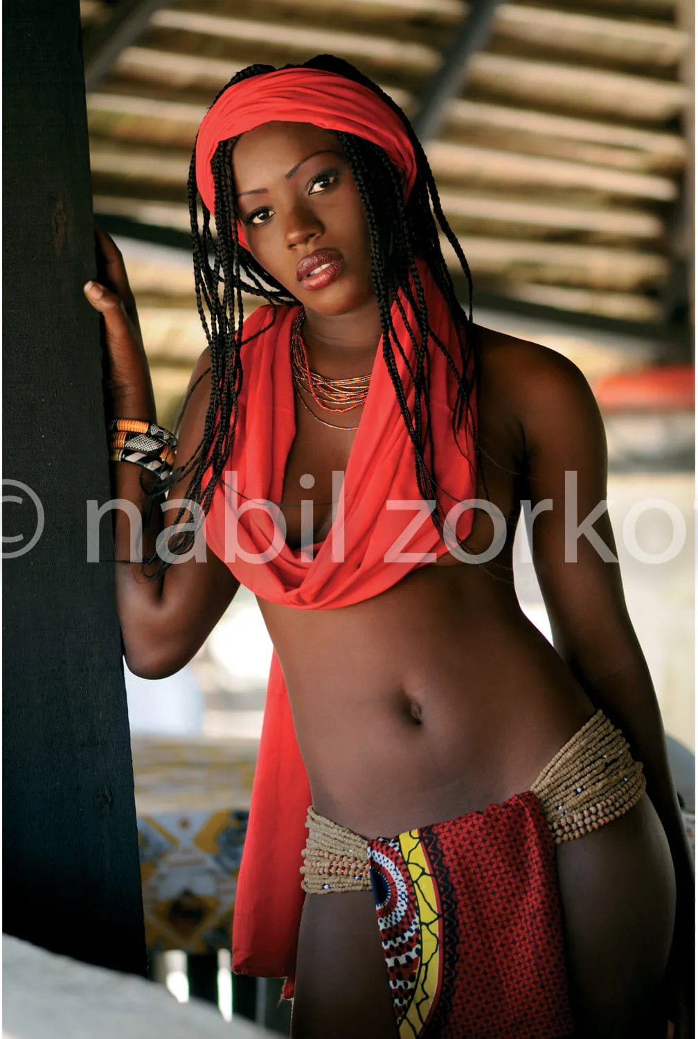 Africa photo — Une femme pose près d’une colonne en bois, vêtue d’une robe rouge drapée, avec un turban rouge assorti et des tresses longues. Elle por