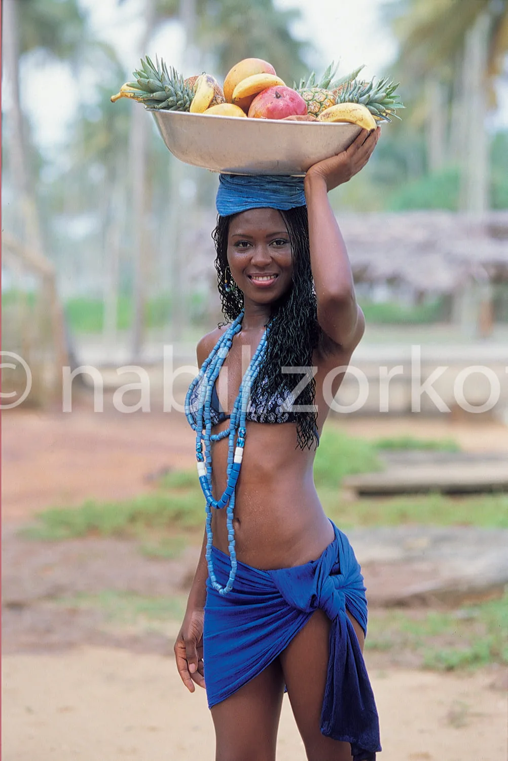 Africa photo — Une femme souriante porte sur la tête un grand panier en bois rempli de fruits tropicaux variés comme des ananas, des mangues et des ba