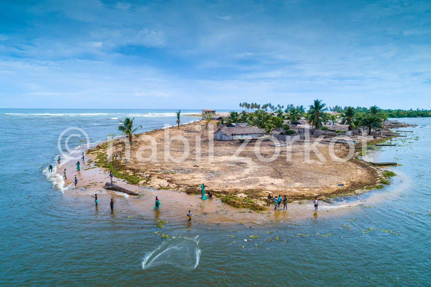 Africa photo — Une vue aérienne d’un petit village côtier entouré d’eau, où des pêcheurs se trouvent sur une étroite bande de sable et dans l’eau peu