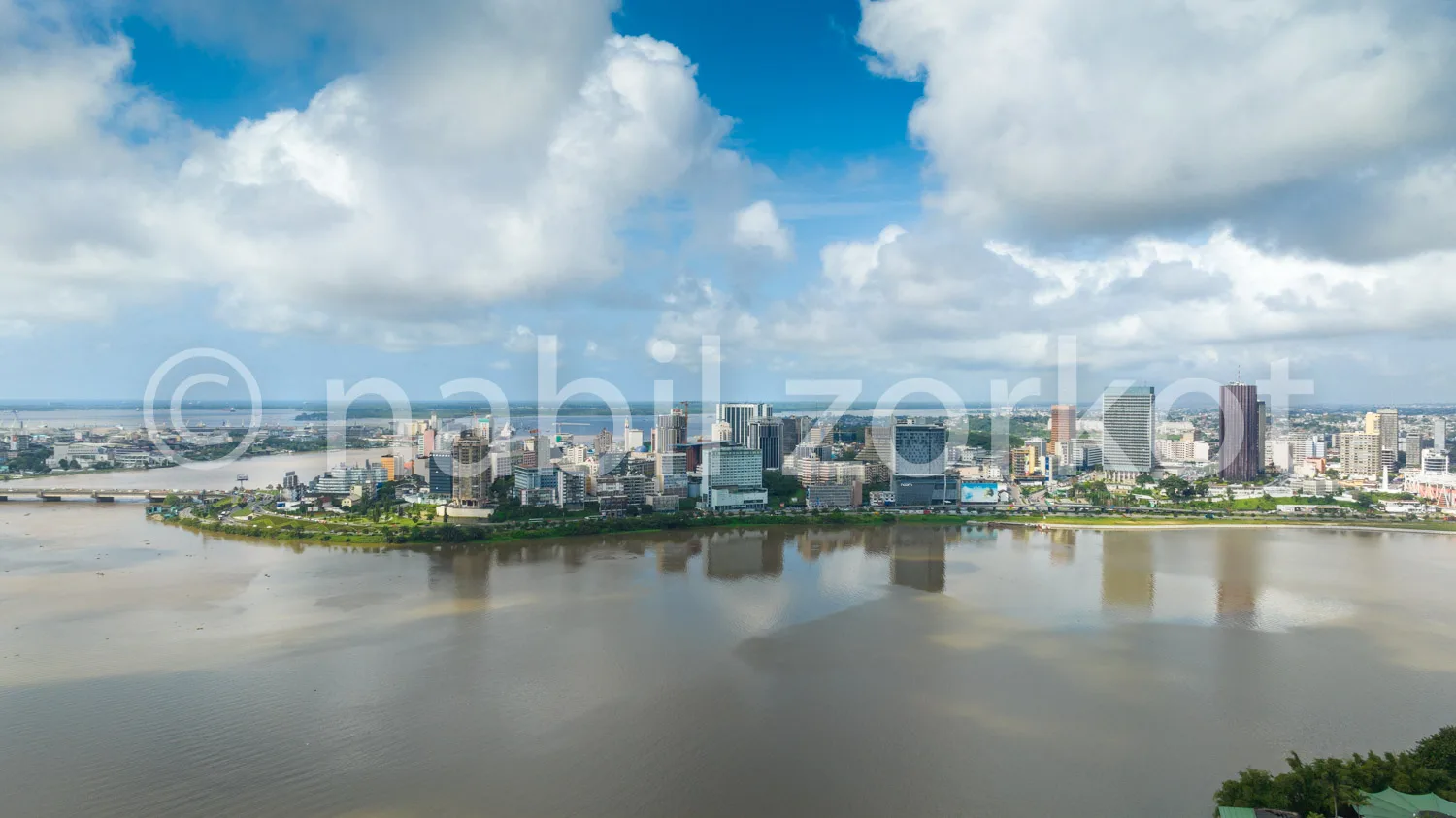 Africa photo — Vue aérienne d'une ville moderne bordée par un large fleuve aux eaux calmes, avec des gratte-ciels et des bâtiments variés en arrière-p
