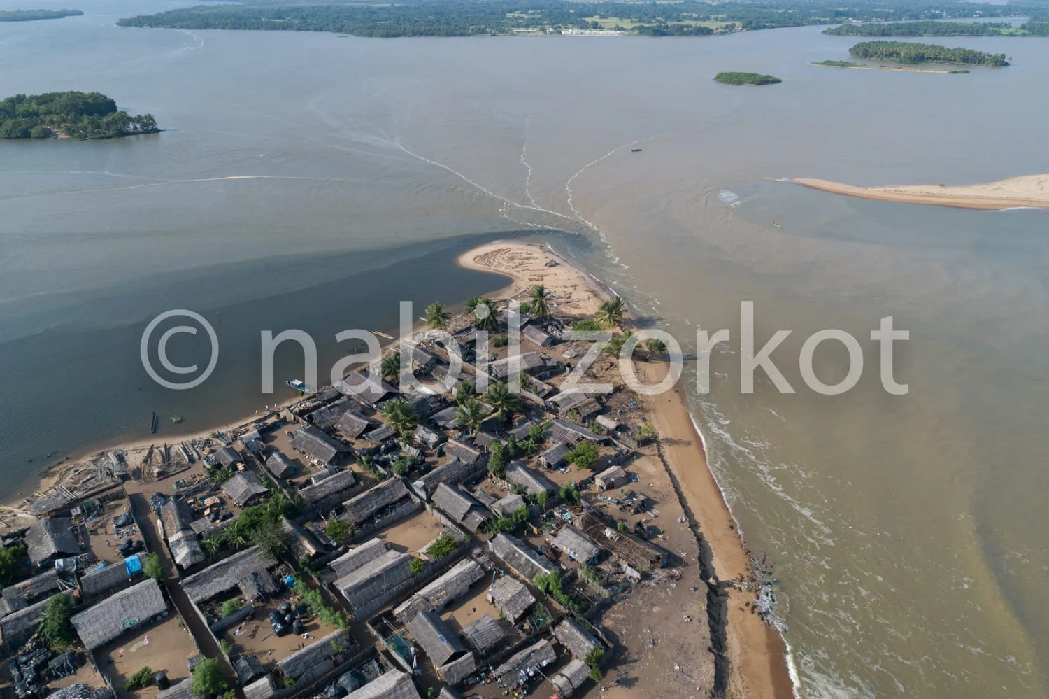 Africa photo — Vue aérienne d’un village côtier composé de maisons en bois aux toits de chaume, bordé par une étroite plage de sable et un large fleuv