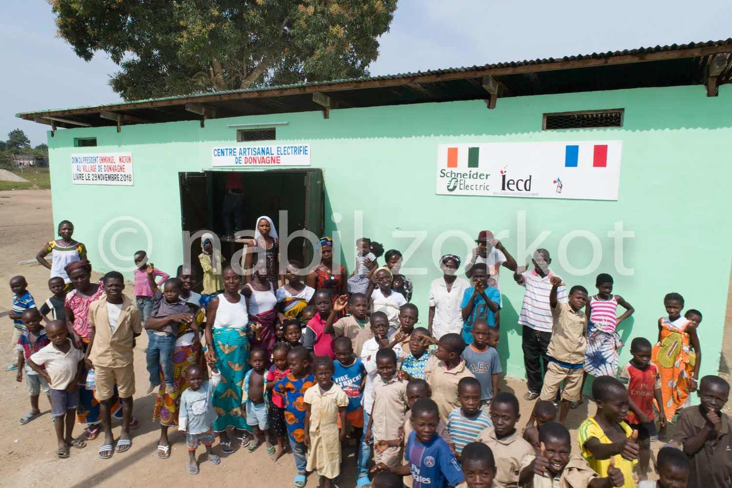 Africa photo — Un groupe joyeux composé d’enfants et d’adultes posant devant un bâtiment vert clair nommé « Centre Artisanal Électrifié de Donvagne ».