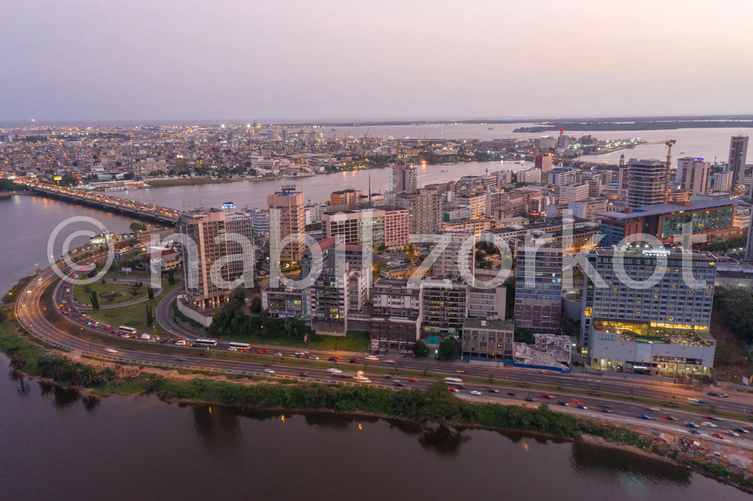 Africa photo — Vue aérienne d’une ville en soirée avec un large fleuve traversant l’horizon, des bâtiments modernes éclairés et une route bien fréquen