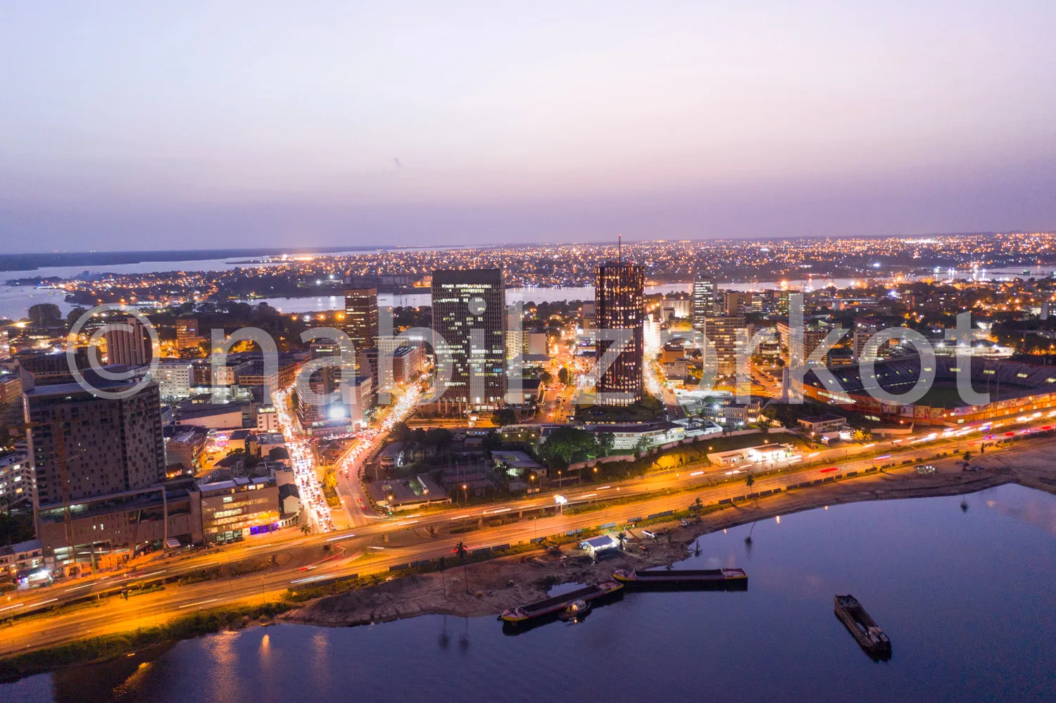 Africa photo — Vue aérienne d’une ville au crépuscule avec des bâtiments éclairés, des routes animées de traînées lumineuses, un fleuve bordé de quais