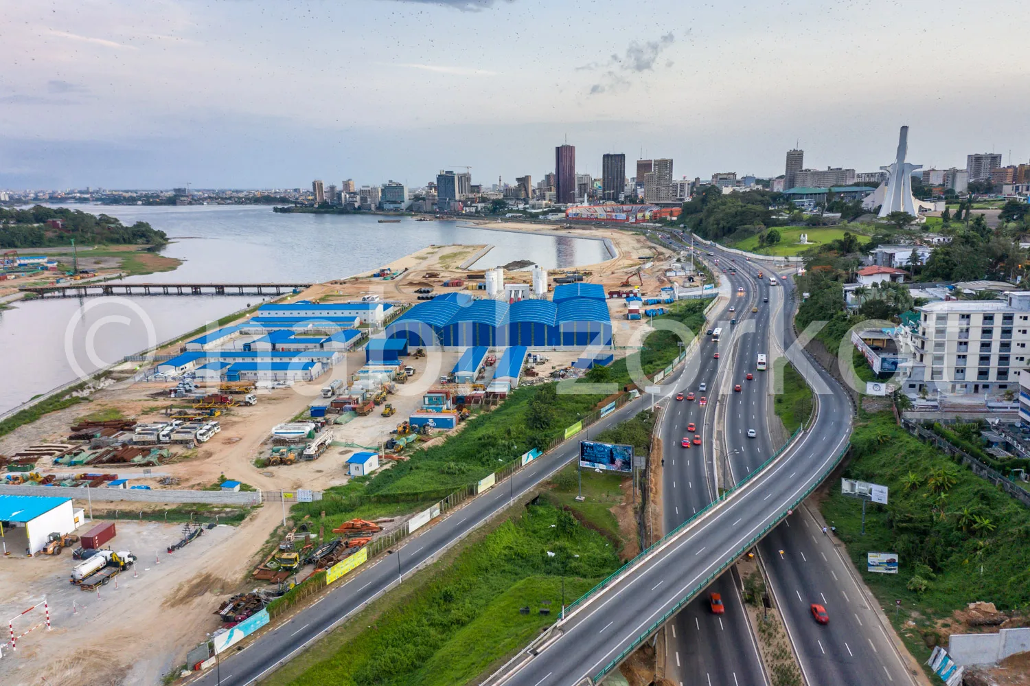 Africa photo — Vue aérienne d'une ville portuaire en développement : une autoroute sinueuse bordée de véhicules, des entrepôts bleus et des chantiers