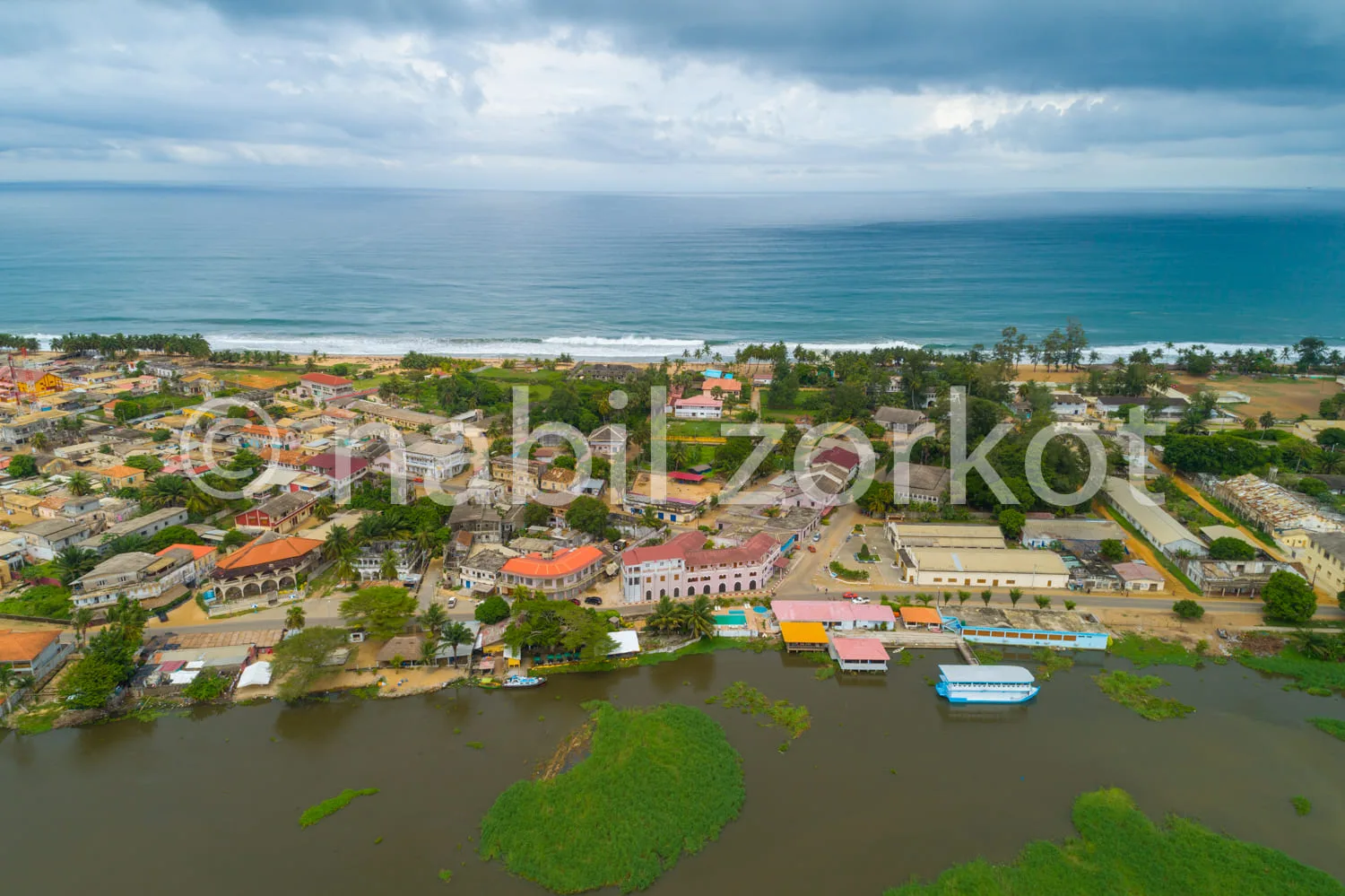 Africa photo — Vue aérienne d’un village côtier avec des maisons colorées, une lagune bordée de végétation et l’océan en arrière-plan sous un ciel nua