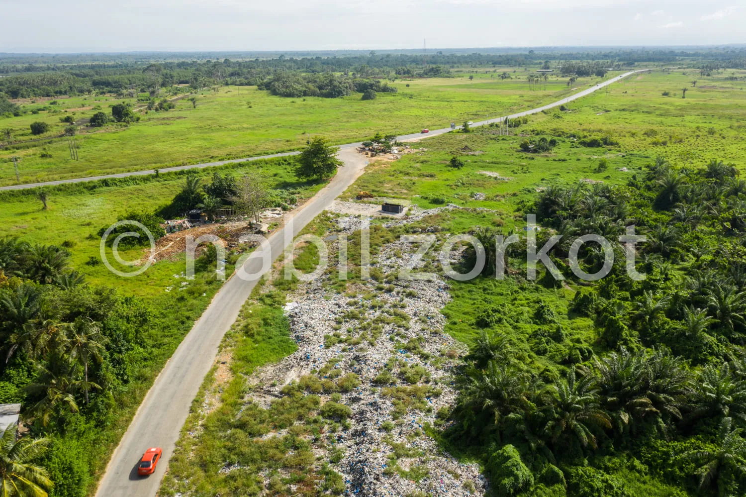 Africa photo — Une route bitumée sinueuse traverse une campagne verdoyante. À gauche, une voiture rouge circule, tandis qu’à droite, un dépotoir sauva