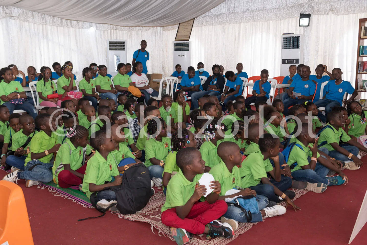 Africa photo — Un groupe d'environ quatre-vingts enfants, vêtus de polos verts, assis en cercle sur des tapis et des chaises dans une salle couverte,