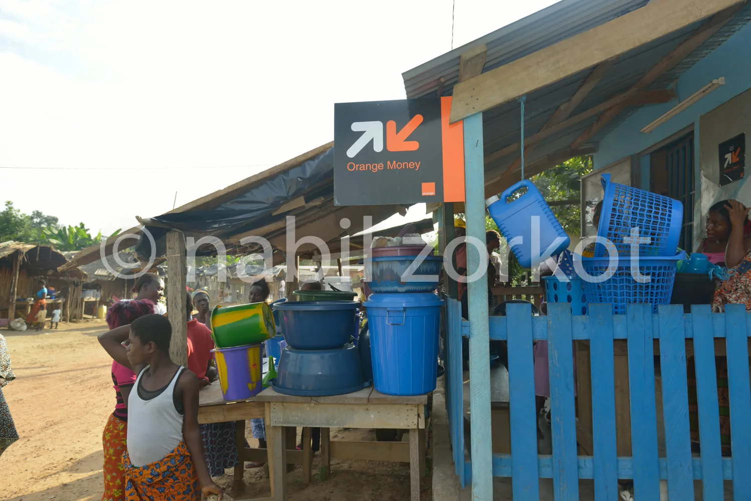 Africa photo — Un point de service Orange Money installé sous une petite structure en bois avec des jerrycans bleus empilés. Une femme en tenue tradit
