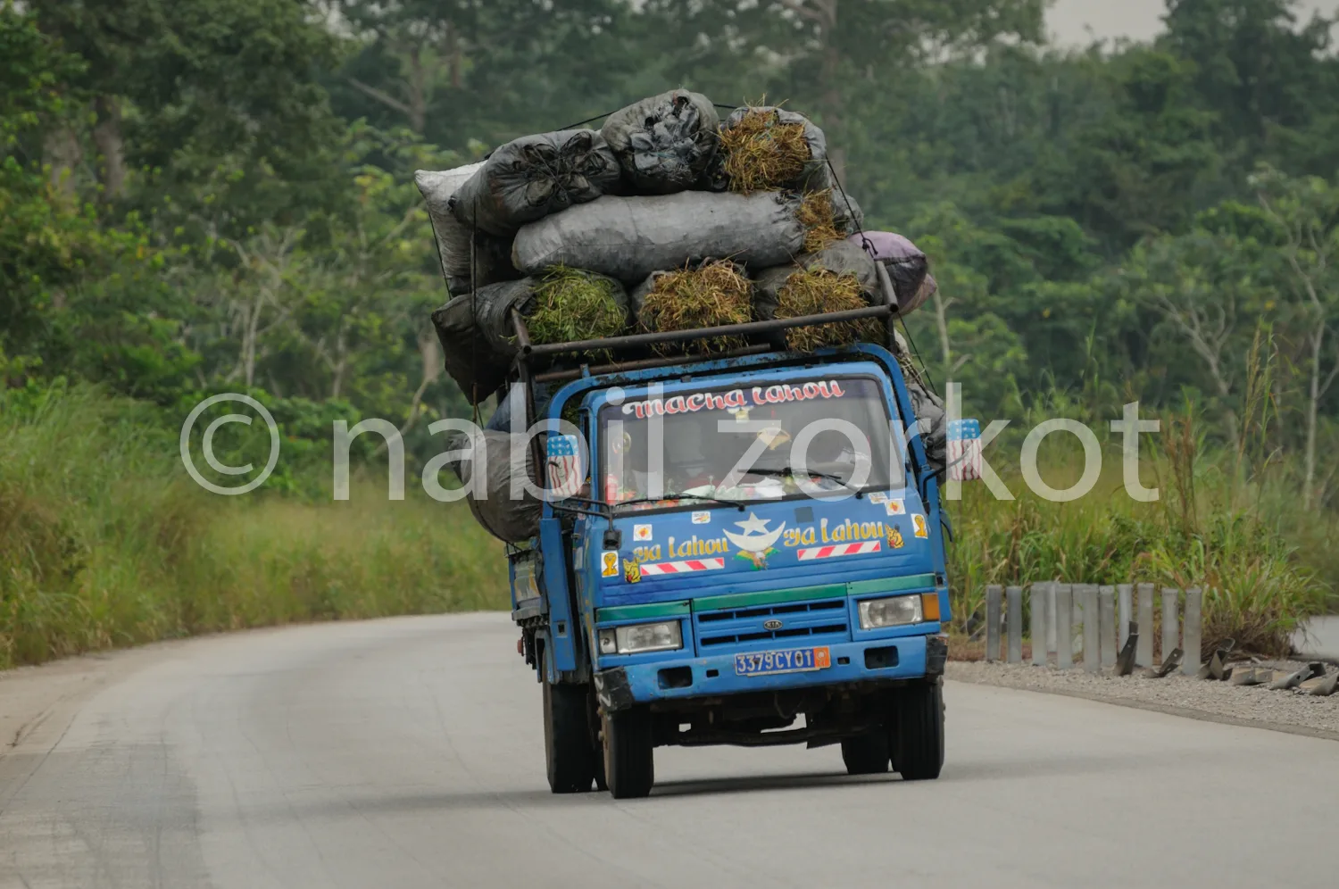 Africa photo — Un camion bleu transportant une charge importante de sacs noirs remplis de charbon végétal, roulant sur une route goudronnée en bordure