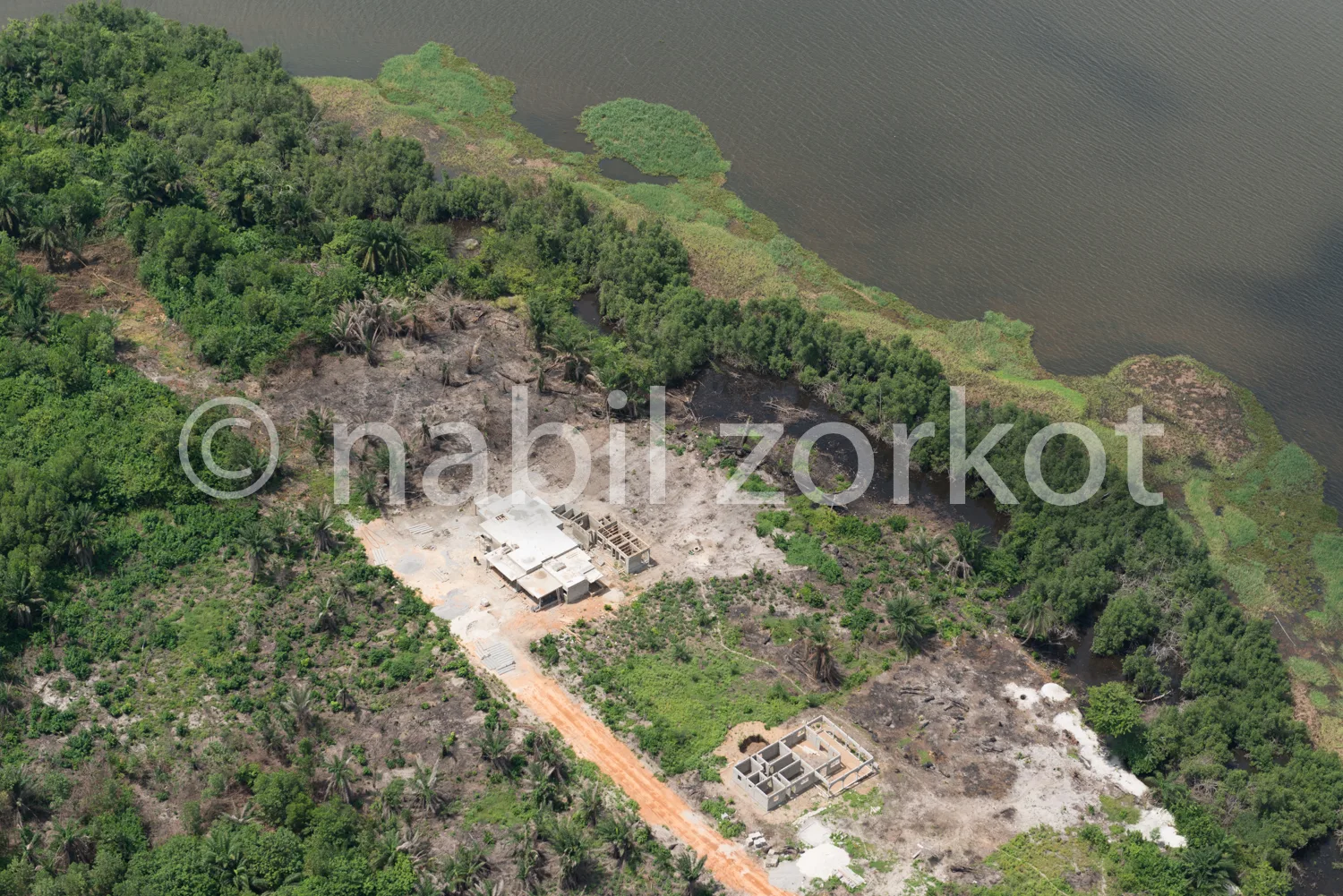 Africa photo — Vue aérienne d’un village isolé entouré de végétation luxuriante, avec deux habitations en bordure d’un lac calme, reliées par un chemi