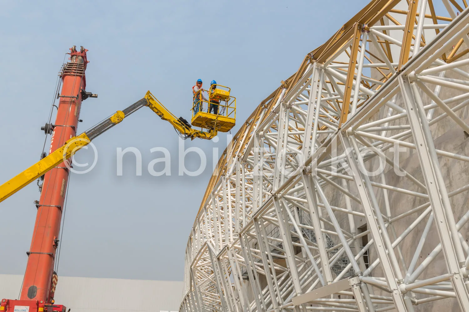 Africa photo — Un ouvrier équipé d’un casque bleu se tient sur une nacelle jaune suspendue à une grue rouge, en train de travailler sur une structure