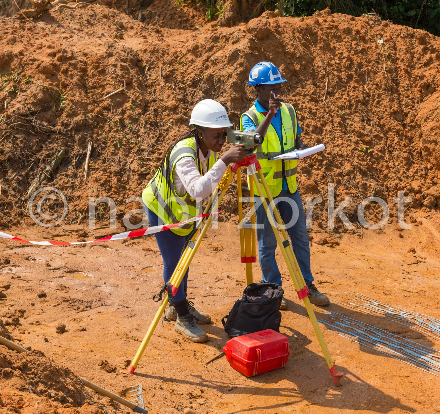 Africa photo — Deux professionnels équipés de casques blancs et gilets jaunes de sécurité utilisent une station totale et un niveau laser sur un terra