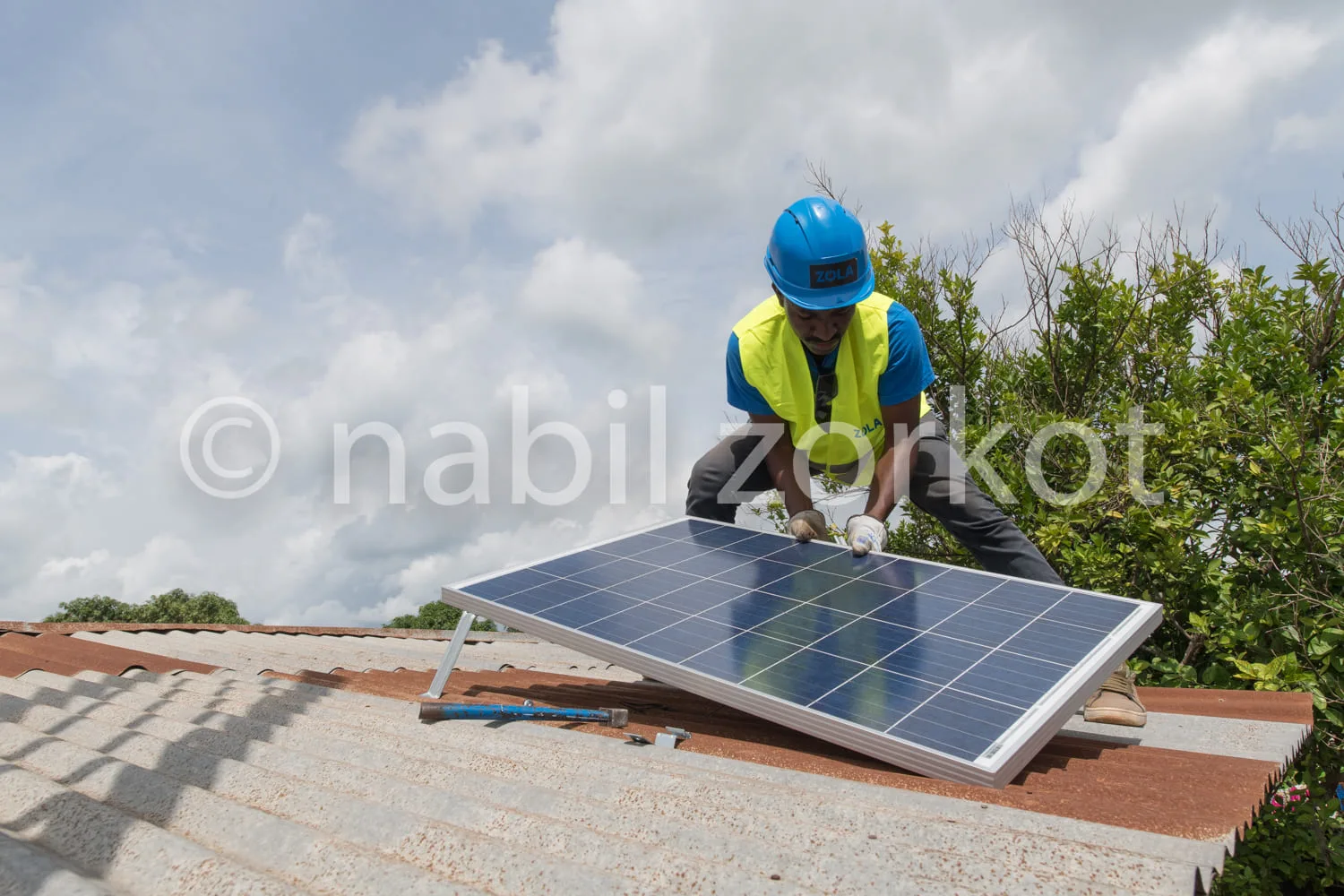 Africa photo — Un professionnel en tenue de sécurité bleue et jaune, casque inclus, fixe un panneau solaire sur un toit en tuiles, sous un ciel dégagé