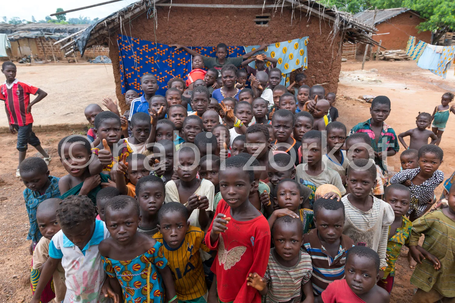 Africa photo — Un groupe d'environ quarante enfants souriants, vêtus de tenues colorées et variées, posent devant une cabane en terre avec une bâche b