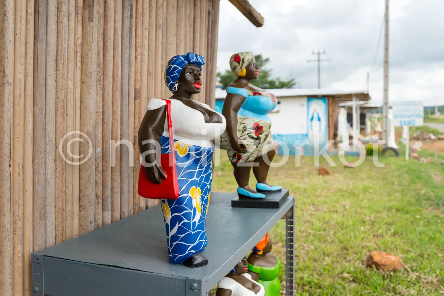 Africa photo — Deux statues en pied représentant des femmes vêtues de tenues traditionnelles colorées, l’une avec un pagne bleu à motifs et un chapeau