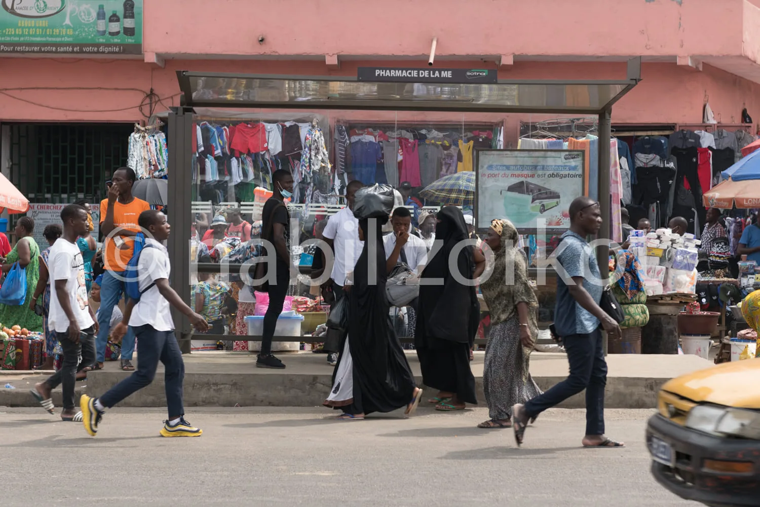 Africa photo — Une pharmacie nommée 'Pharmacie de la Me' située dans un marché animé. Des vêtements de différentes couleurs sont suspendus à l'extérie