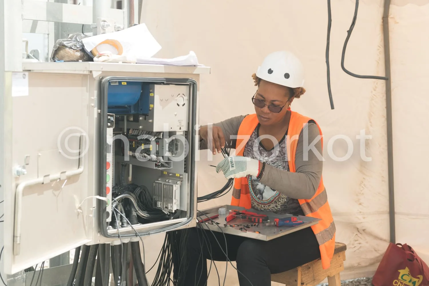 Africa photo — Une technicienne équipée d’un casque de protection blanc et d’un gilet orange examine attentivement un tableau électrique ouvert, rempl