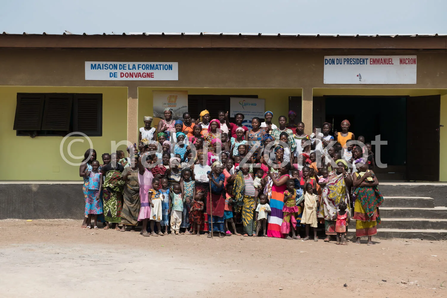 Africa photo — Un groupe de personnes, majoritairement des femmes vêtues de tenues colorées traditionnelles, posent souriantes devant la Maison de la