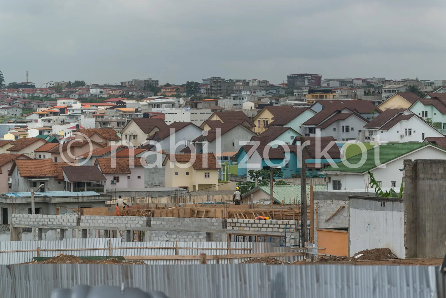 Africa photo — Vue d’un chantier de construction en cours avec des fondations en béton, des murs de bloc et des échafaudages, entouré de maisons indiv
