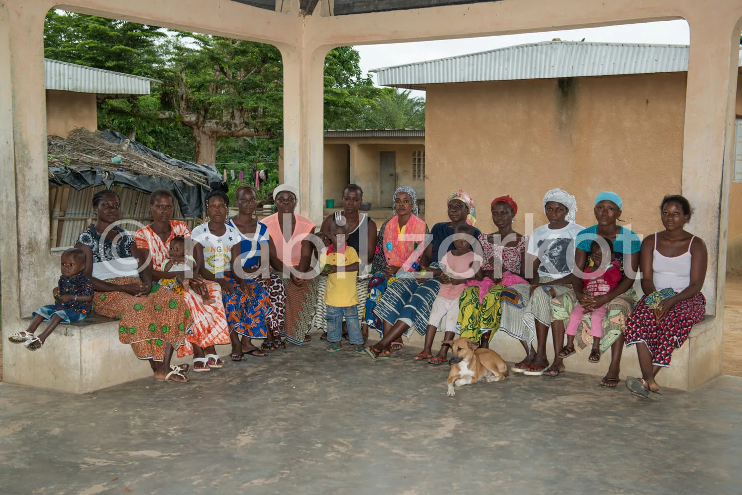Africa photo — Un groupe de douze femmes assises sur un muret bas, souriantes, vêtues de pagnes aux motifs variés et de coiffures traditionnelles. Un