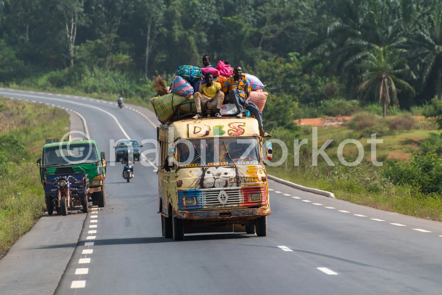 Africa photo — Un camion rempli de passagers assis sur le toit et à l'intérieur, roulant sur une route bitumée entourée de végétation tropicale. Le vé