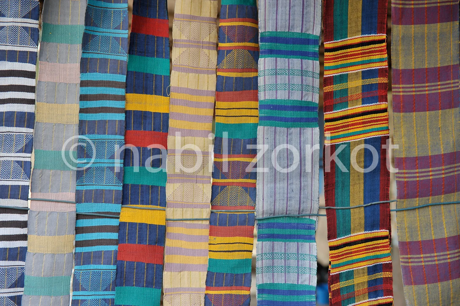 Africa photo — Une série de tissus artisanaux suspendus verticalement, présentant des motifs géométriques variés en bandes horizontales de couleurs vi