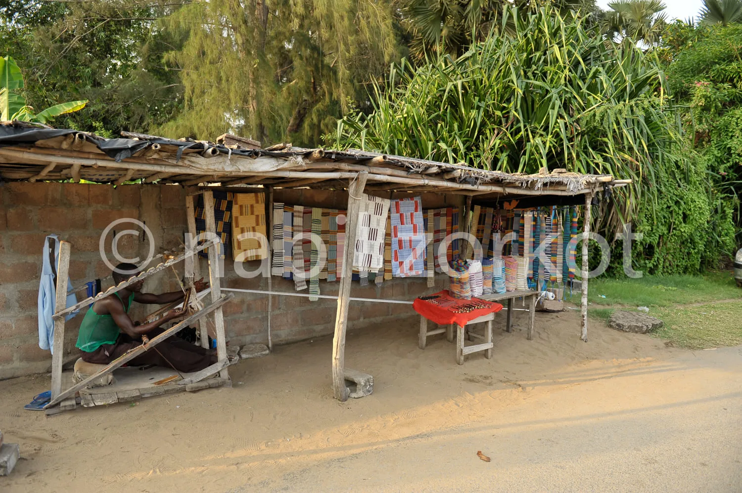 Africa photo — Un homme est assis sous un abri de fortune en bois et tôle, entouré de tissus colorés suspendus et étalés sur des tables, dans un cadre