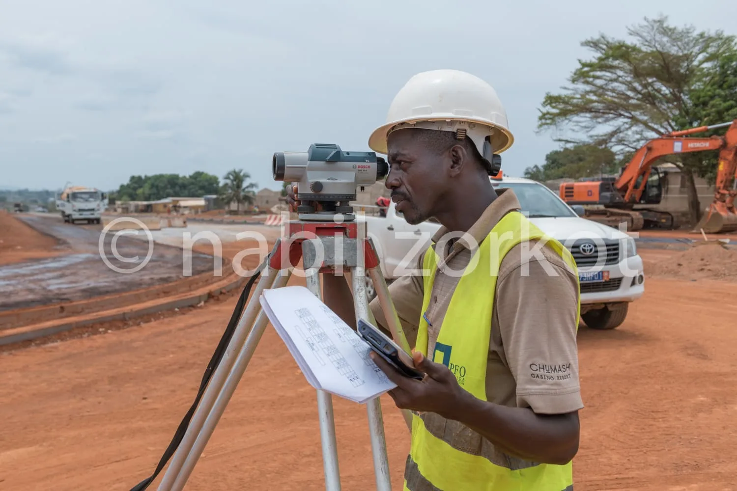 Africa photo — Un professionnel en tenue de sécurité, casque blanc et gilet jaune, utilise un théodolite de précision pour effectuer des relevés sur u