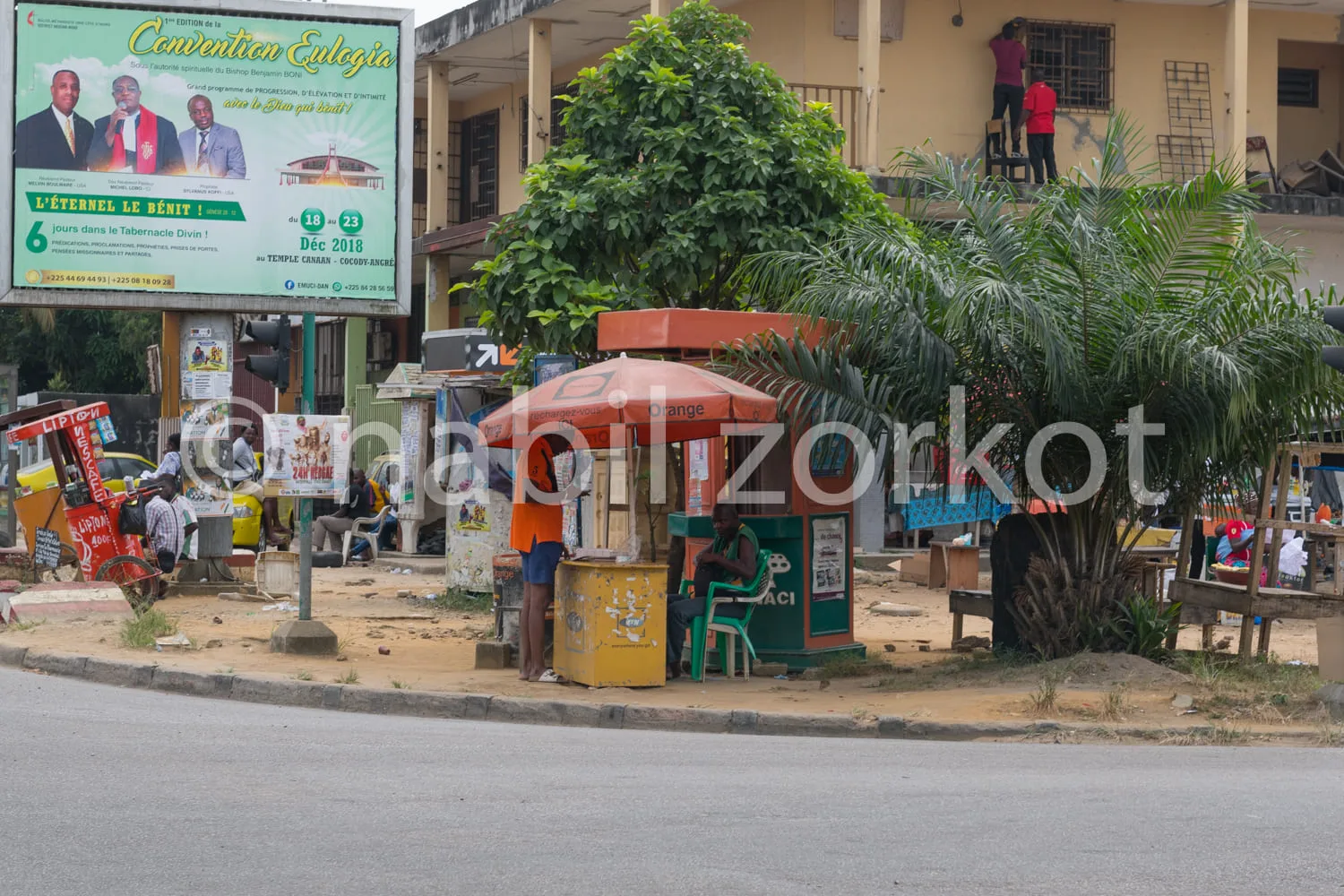 Africa photo — Un kiosque à journaux coloré avec un parasol orange est installé sur un trottoir. À l’arrière-plan, une grande affiche annonce une conv