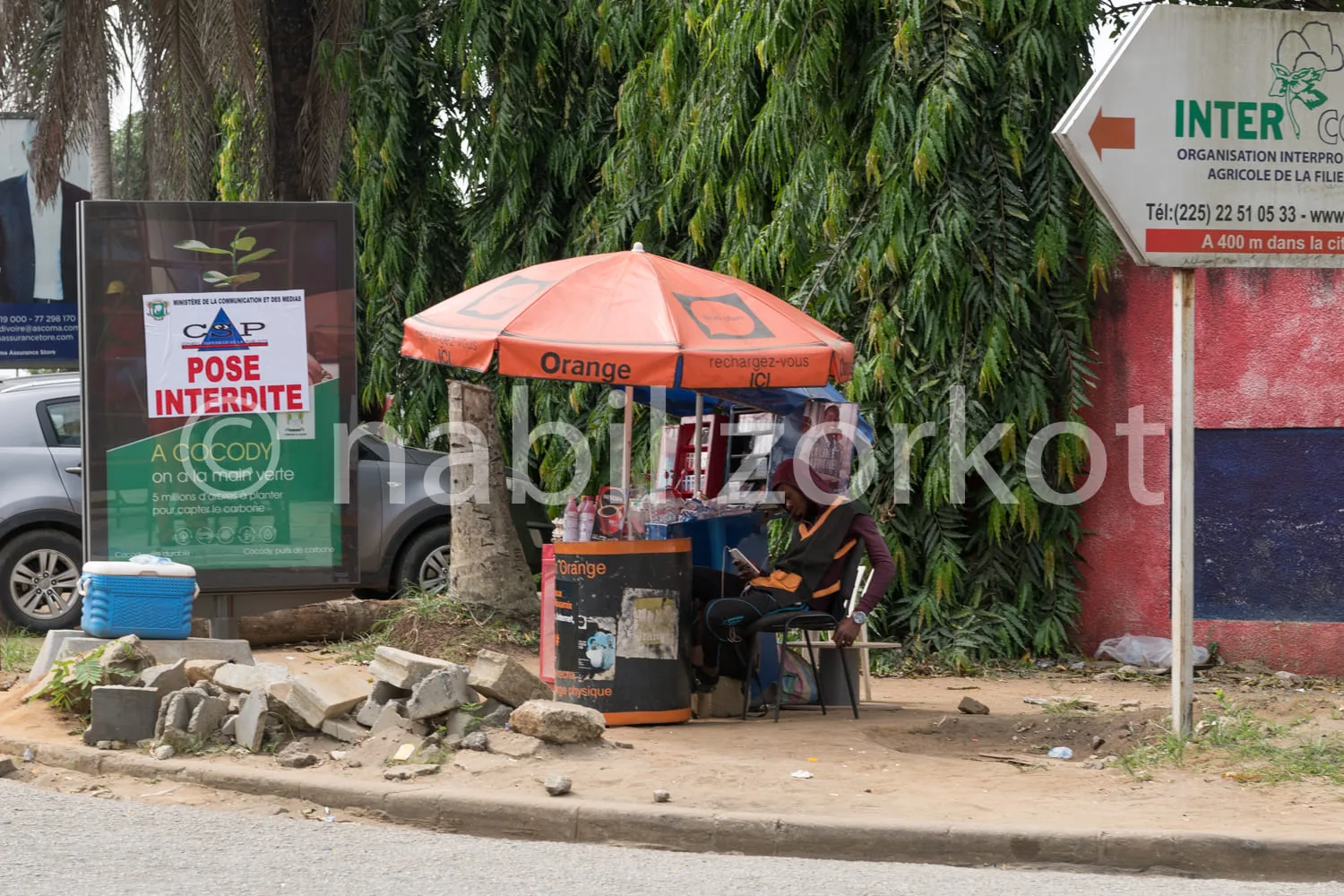 Africa photo — Un commerçant assis sous un parasol orange, près d’un petit kiosque de recharge de téléphones mobiles. À côté, une affiche indique une